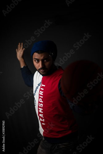 Fashion portrait of an young Indian Bengali brunette man in casual tee shirt and head band playing with basket ball in black copy space background. Indian lifestyle and fashion photography.