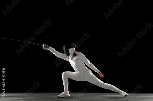 Catching moment. Teen girl in fencing costume with sword in hand isolated on black background. Young female model practicing and training in motion, action. Copyspace. Sport, youth, healthy lifestyle.