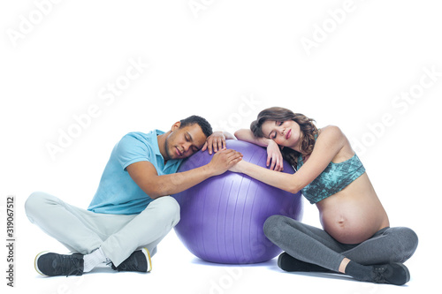 A young couple and their wife in the last months of pregnancy are sitting on a sports mat near the ball. Black man and white woman. The concept of a happy couple of different races preparing together