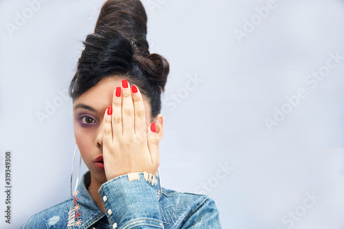 Serious young girl looking with one eye at the camera as she covers the other with her hand in a close up portrait.