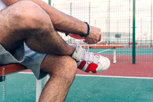 young guy in shorts on the sports ground tying shoelaces on his beautiful sneakers while sitting on a bench on a warm summer day