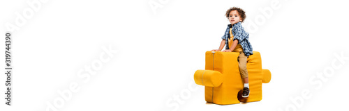 panoramic shot of little african american boy sitting on yellow puzzle chair, isolated on white