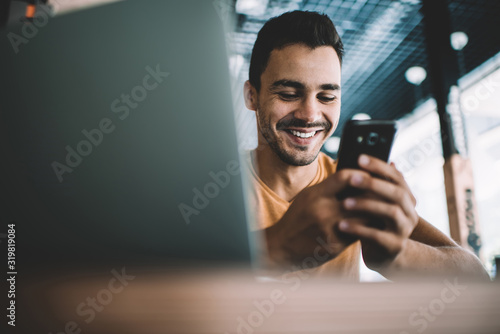  Millennial male laughing watching funny video on smartphone spending free time in cafe interior,successful man blogger satisfied with online earning getting income on banking account on cellular.