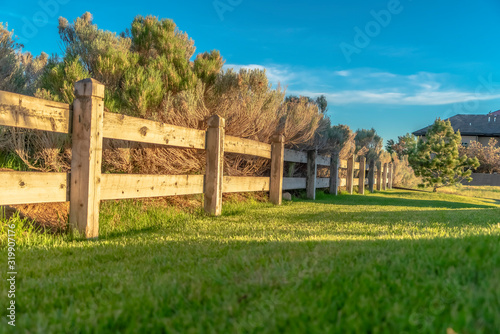 Brown wooden low fence on a lush green field against thick bushes and shrubs