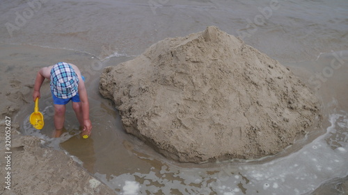 Dziecko na plaży bawi się w piasku nad morzem. Chłopiec z wiaderkiem i łopatką buduje zamki z piasku. Ma założoną czapeczkę przeciwsłoneczną. Dobrze się bawi.