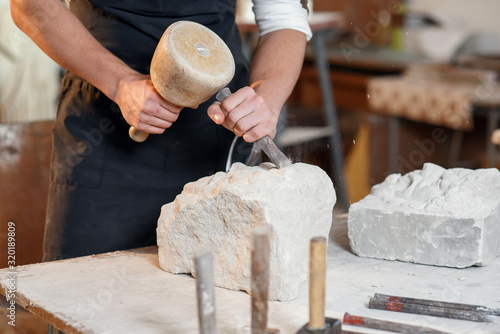 Bearded craftsman works in white stone carving with a chisel. Creative workshop with works of art.