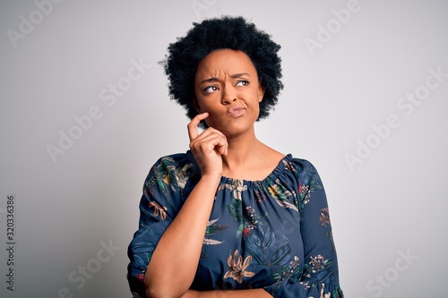 Young beautiful African American afro woman with curly hair wearing casual floral dress with hand on chin thinking about question, pensive expression. Smiling with thoughtful face. Doubt concept.