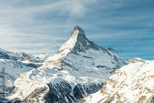 Scenic view on snowy Matterhorn (Cervin, Cervino) peak in sunny day with blue sky, pine trees, valley and clouds in background. Panoramic landscape in white and blue colors in Zermatt, Switzerland