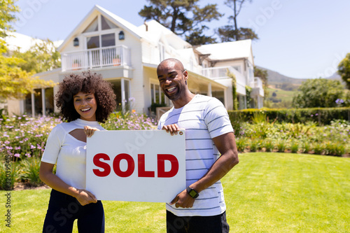 Happy young african american couple in the garden holding sold sign