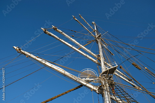 The main mast and yards of a romantic classic sailing ship, visible shrouds, stays, mast nest, and complex rigging, blue sky in the background.