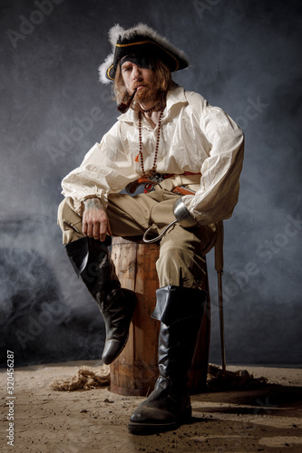 Pirate filibuster sea robber in suit with gun and saber sits on barrel. Concept photo