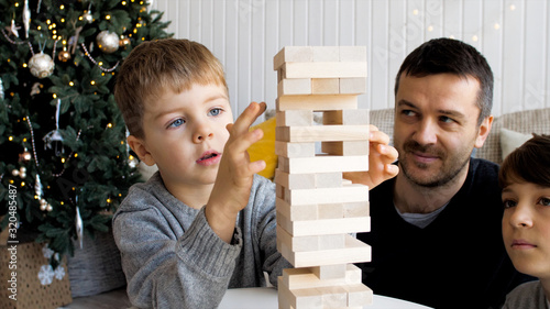 Family is playing in board game with wooden tower together at home. Junior son is pulls out a brick while older brother and father is looking on it.