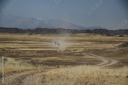 samochody terenowe w oddali na polnej drodze pomiędzy wyschniętymi trawami afrykańskiej sawanny z górami w tle
