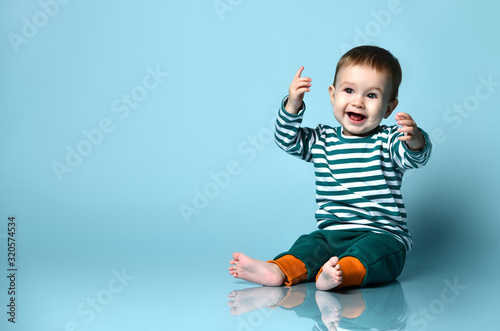 Little baby boy in stylish casual clothing barefoot sitting on floor and smiling with raised hands over blue wall background