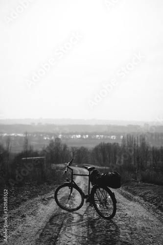 Outdoor view on bicycle with things, a tent on the trunk.. hilltop overlooking a valley in haze, a city on the horizon. winter or autumn landscape dirt road. vertical photo black and white