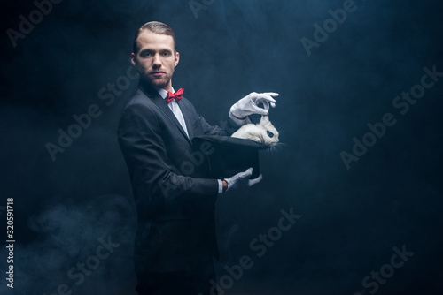 professional magician in suit showing trick with white rabbit in hat, dark room with smoke