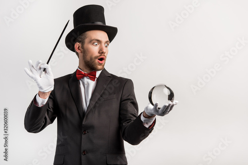 shocked magician holding wand and magic ball, isolated on grey