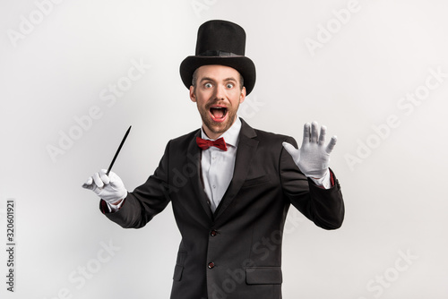 excited magician in suit and hat holding wand, isolated on grey