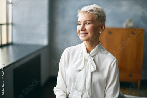 Attractive middle aged lady in stylish white silky blouse thinking about something pleasant, sitting by window in office interior, rejoicing at good day, ready to do work routine. People and lifestlye