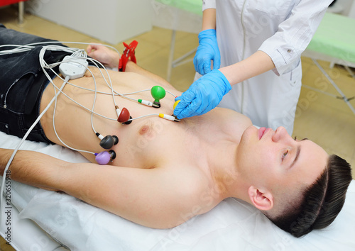 a young male patient in a modern clinic checks the state of the heart and makes an ECG or electrocardiogram with vacuum rubber colored sensors electrodes on the chest.