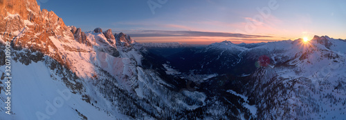 Aerial, panoramic winter view on Pale di San Martino mountains and valley covered in snow. Vezzana, Dolomites. Red rocks lit by setting sun against blue sky. San Martino di Castrozza, Italy.