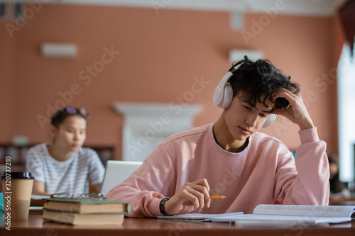 Pensive teenage student with headphones looking through page of manual before rewriting information in his notebook