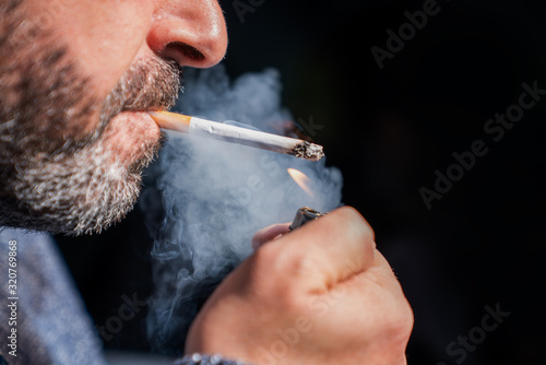 Close up of bearded man holding a lighter and smoking a cigarette on black background.