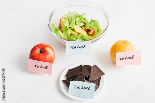 Fresh fruits, chocolate and salad with calories on cards on white background, calorie counting diet