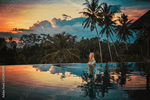 Woman relaxes in a luxury infinity pool overlooking the jungle at sunset in Ubud, Bali. A girl sits on the edge of the infinity pool against the backdrop of a bright beautiful sunset and the jungle.