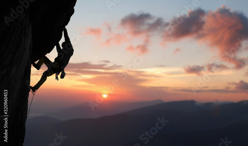 Male climber in rock climbing equipment, pulling up and doing next step on high cliff reaching rocky top. Side view. Panoramic view of mountains and amazing pink sky with sunset and clouds. Copy space