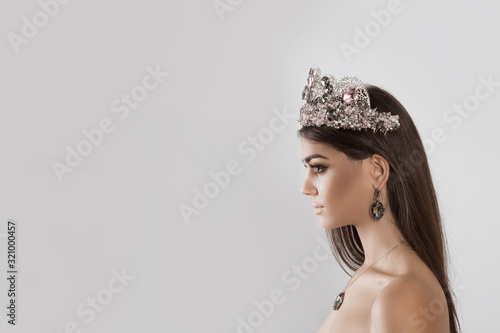 Closeup portrait in profile Beauty queen pageant winner girl with tiara long hair lipstick and eye shadows matching with dark crystal diamond earring necklace and crown looking to side on white grey