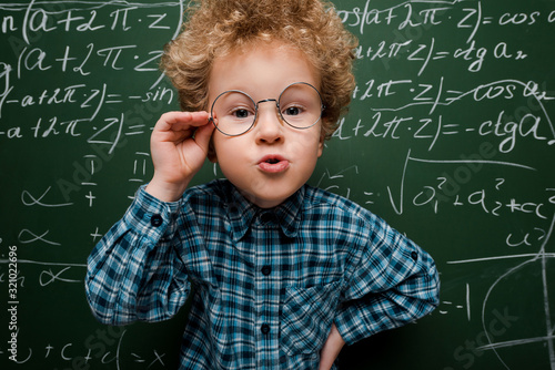 smart kid touching glasses and standing with hand on hip near chalkboard