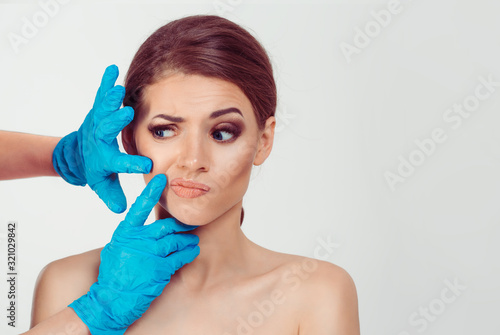 Preparing for cosmetic plastic surgery . Skeptical worried woman looks to the surgeon hands while the doctor examines her nasolabial wrinkle crease doubtful scared afraid, isolated white background