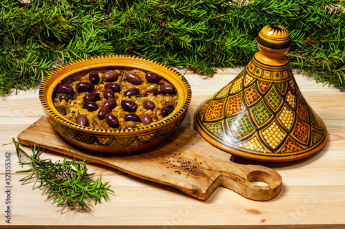 Moroccan tagine dish in open traditional tagine pot with black olives on the top. Tagine placed on wooden (olive wood) board. Common yew foliage at the background.