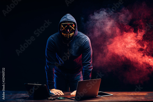 young programmer in a smoke-filled background, a hacker in mask, trying to hack into a computer system, unrecognizable male stand above table with laptop