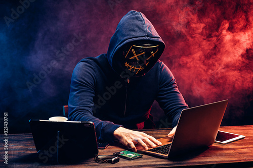 hacker man typing on laptop, hacking computer system. male in mask and pullover. unrecognizable incognito male sit in hood and try to breach the security of laptop system. neon smoke in background