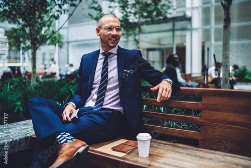 Caucasian professor in luxury elegant clothing and bifocal spectacles resting at urban bench in metropolitan downtown, formally dressed adult student 30 years old holding cellphone in hand and smiling