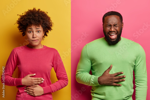 Horizontal shot of dark skinned couple touch stomachs, suffer from chronic gastritis, being hungry has displeased face expressions, isolated over yellow and pink background. Healthcare concept