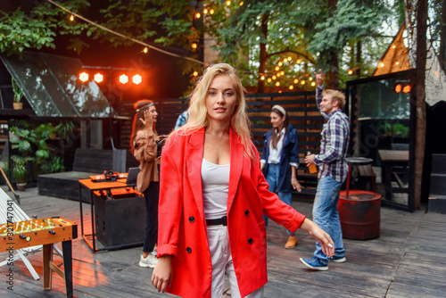 Close up of attractive blond girl with blue eyes posing to the camera with her friends dancing in disco light on the background. Outdoors evening party concept.