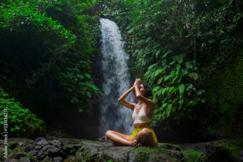 outdoors portrait of young attractive and happy hipster girl doing yoga at beautiful tropical waterfall meditating enjoying freedom and¡ nature in wellness and zen lifestyle