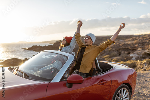 Joyful couple enjoying vacations, driving together convertible car on the rocky ocean coast on a sunset. Happy vacation, love and travel concept