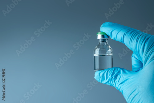 Hand in a blue glove holds a bottle with a vaccine close-up on a gray background, medical concept vaccination subcutaneous injection, dose. Disease treatment immunization.