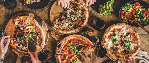 Family or friends having pizza party dinner. Flat-lay of people eating various kinds of Italian pizza and drinking wine over wooden table, top view, wide composition. Fast food lunch concept