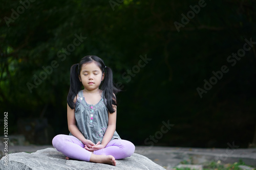 Asian child cute or kid girl close eye mindfulness meditating with breath and sit on stone or rock for peace and relax or yoga on nature green garden and tree at temple park or meditate church outdoor