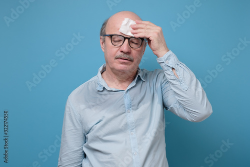 Senior hispanic man with sweat stain on his shirt suffering from hot weather. Studio shot on blue wall. Problem with hyperhidrosis