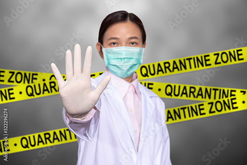 Asian female doctor wear a medical mask, raise hand for stop sign to do not enter quarantine area with yellow quarantine sign on the back, entrance is forbidden in quarantine zone.