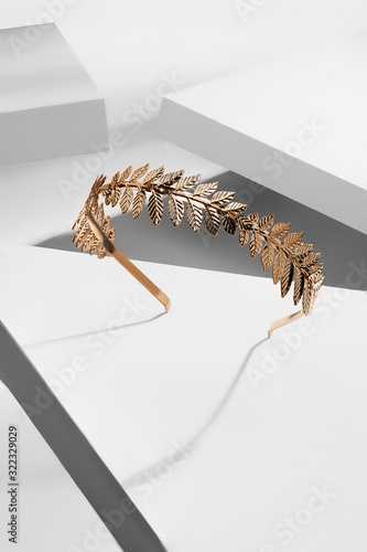 Subject shot of a metal headband made as a crown of golden embossed leaves. The fancy headband is isolated on the white surface surrounded with geometric details.