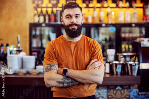 Young handsome bearded tattooed cafe owner standing with arms crossed and looking at camera.