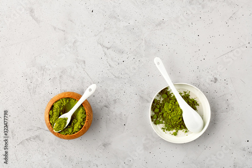 Matcha green tea powder in oak cup with white ceramic spoon and in ceramic cup with white ceramic spoon.