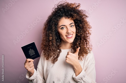 Beautiful tourist woman with curly hair and piercing holding canada canadian passport id happy with big smile doing ok sign, thumb up with fingers, excellent sign